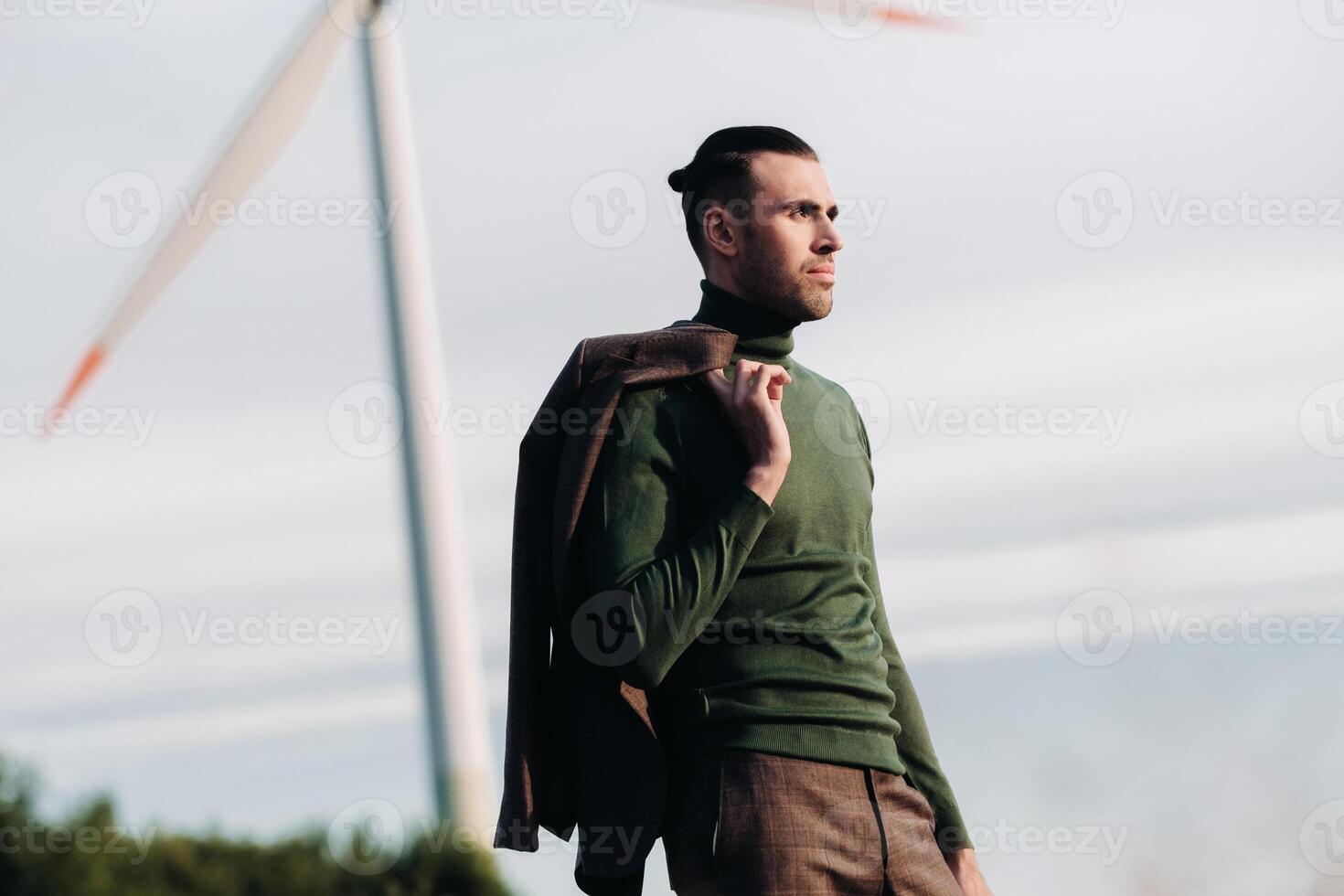 A man in a business suit with a green Golf shirt stands next to a windmill against the background of the field and the blue sky.Businessman near the windmills.Modern concept of the future. photo