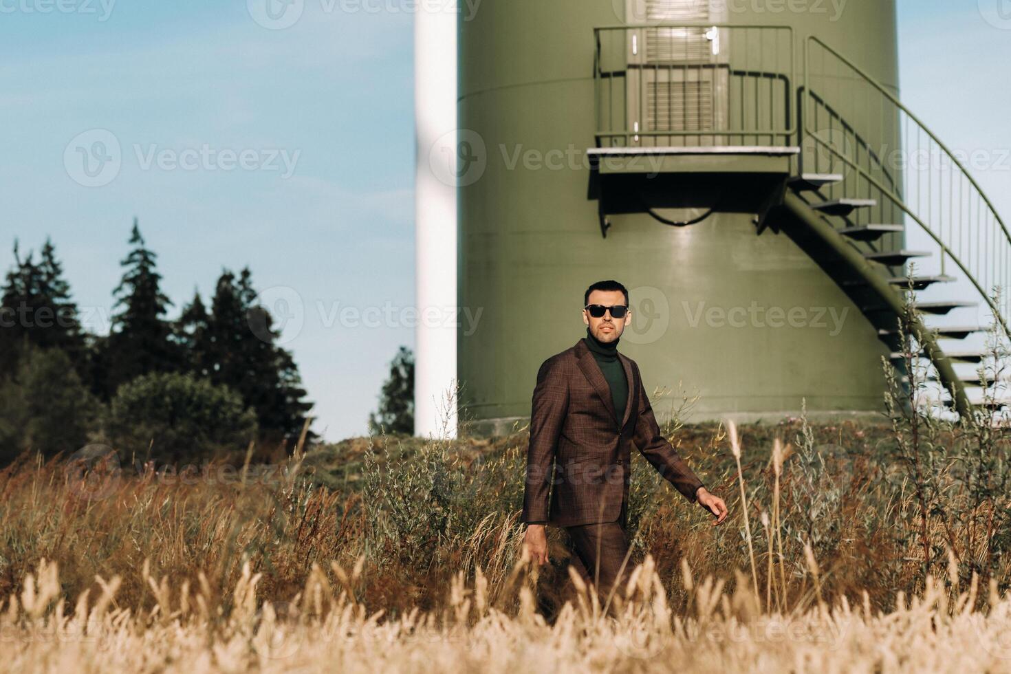 A man in a business suit with a green Golf shirt stands next to a windmill against the background of the field and the blue sky.Businessman near the windmills.Modern concept of the future. photo