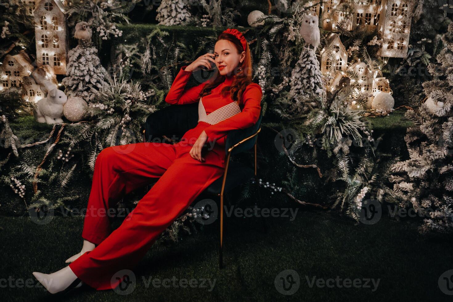 a girl in a red suit is sitting on a chair near the Christmas background. a woman in red on the background of Christmas trees and small Christmas houses photo