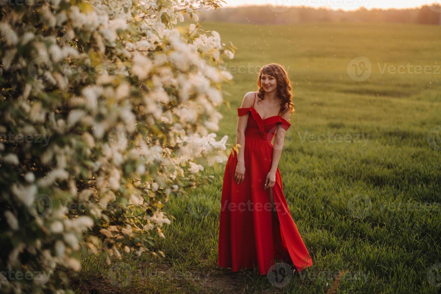 a girl in a red dress with red lips stands next to a large white flowering tree At sunset photo
