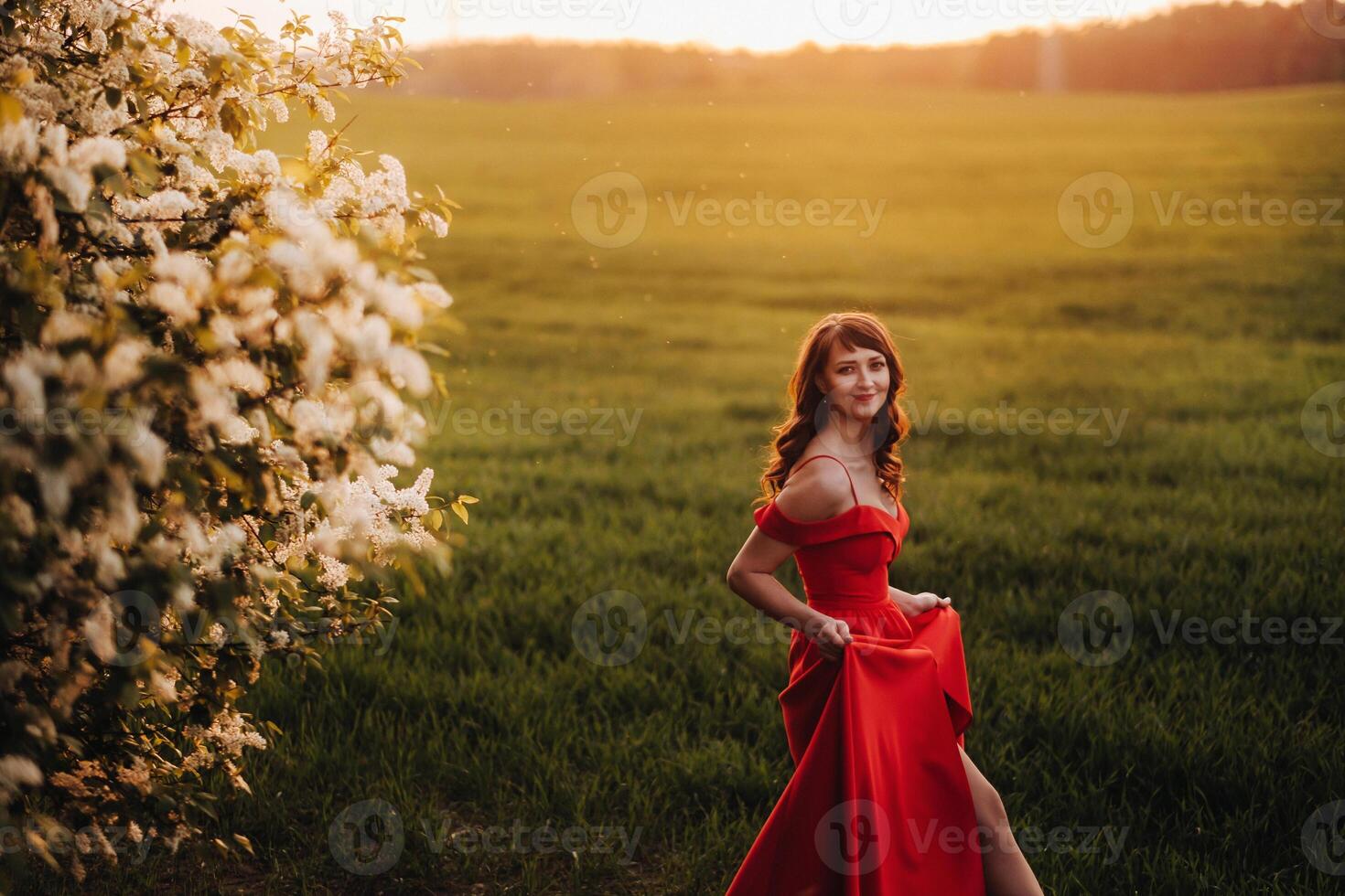 a girl in a red dress with red lips stands next to a large white flowering tree At sunset photo