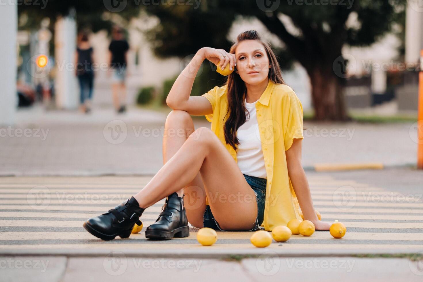a girl with lemons in a yellow shirt, shorts and black shoes sits on a yellow pedestrian crossing in the city. The lemon mood photo