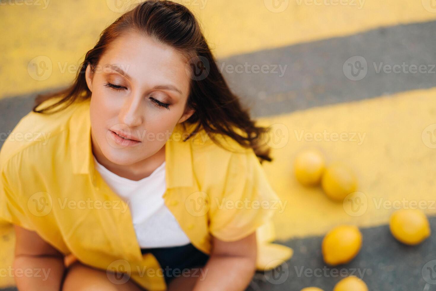 a girl with lemons in a yellow shirt, shorts and black shoes sits on a yellow pedestrian crossing in the city. The lemon mood photo