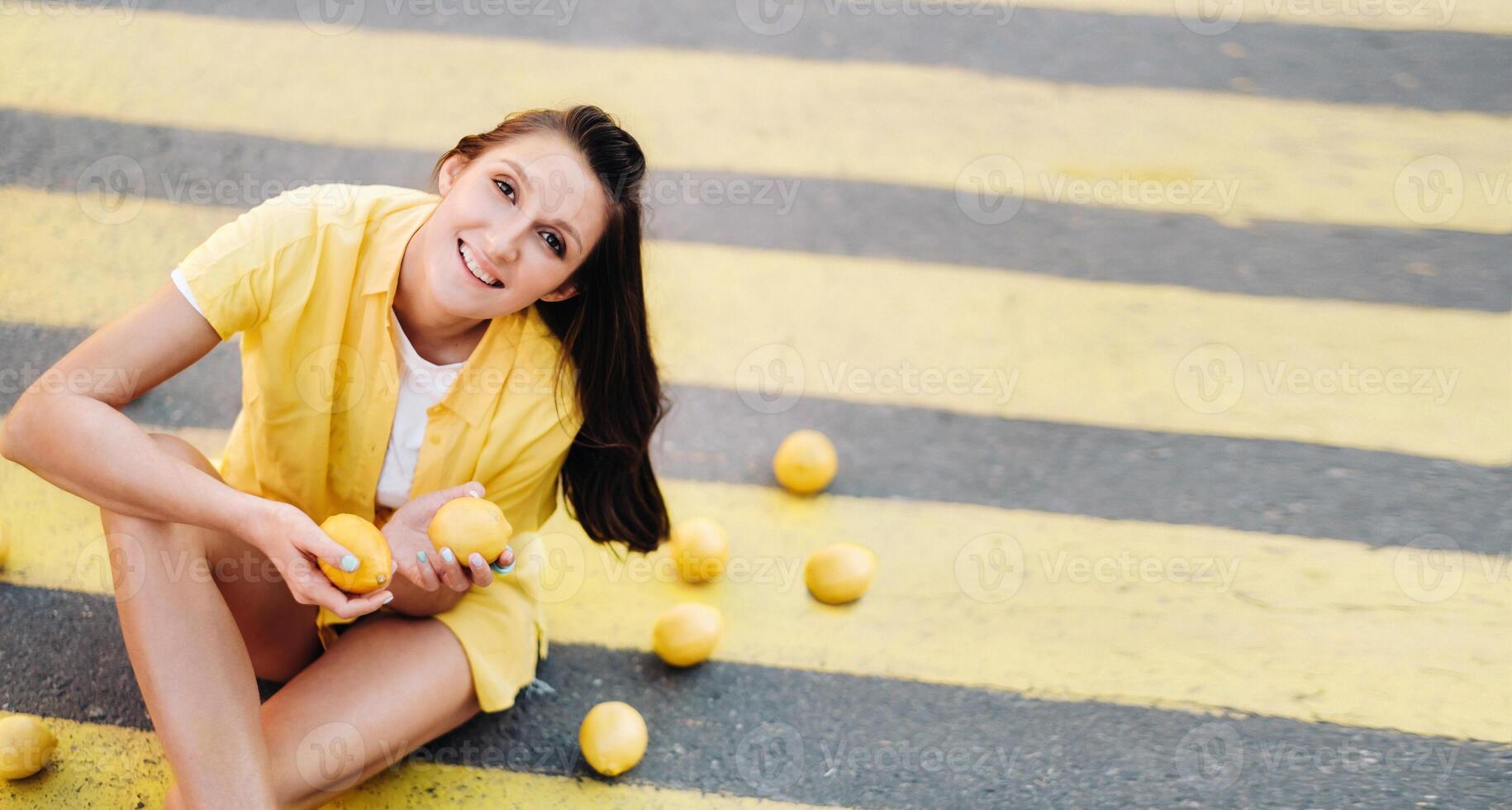 a girl holding lemons in her hands dressed in a yellow shirt, shorts and sitting on a yellow pedestrian crossing in the city and smiling. The lemon mood photo