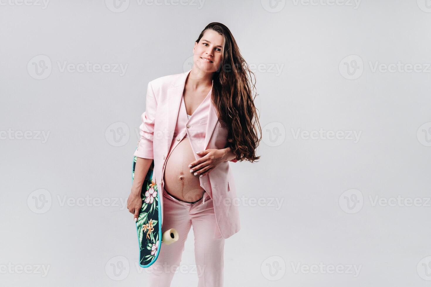 A pregnant girl in a pink suit with a skateboard in her hands stands on a gray background photo