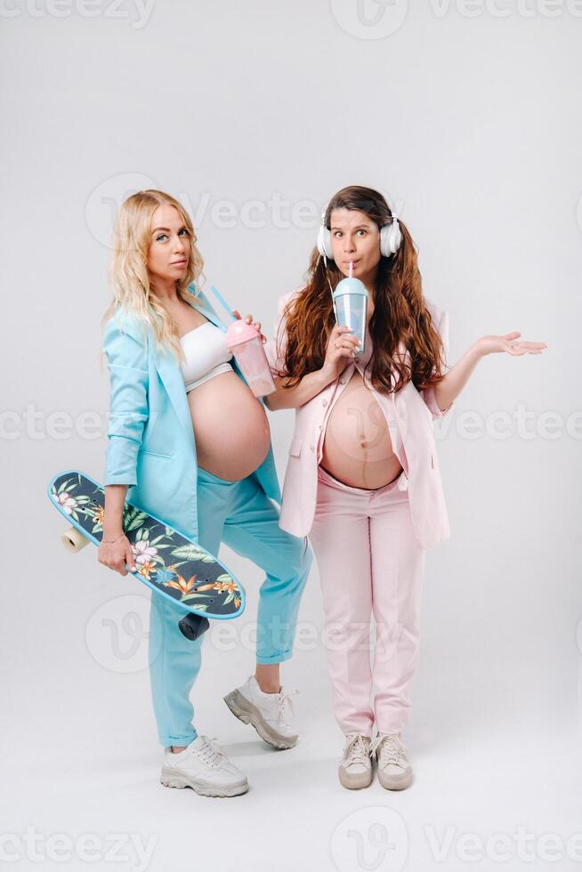 Two pregnant girls in turquoise and pink suits with glasses of juice, a skate and headphones stand on a gray background photo