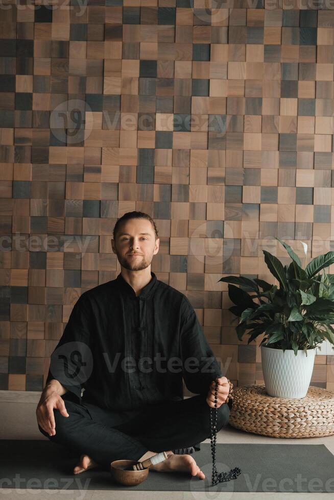 Portrait of a young man in a black kimano sitting in a lotus position on a gym mat in the interior photo