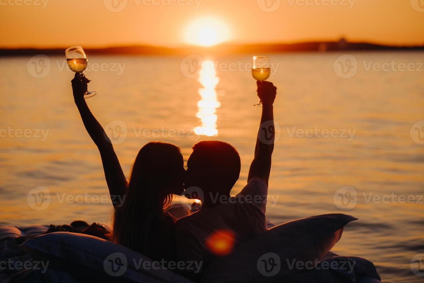 Silhouettes of a happy couple raising glasses on a summer evening near the sea at sunset photo