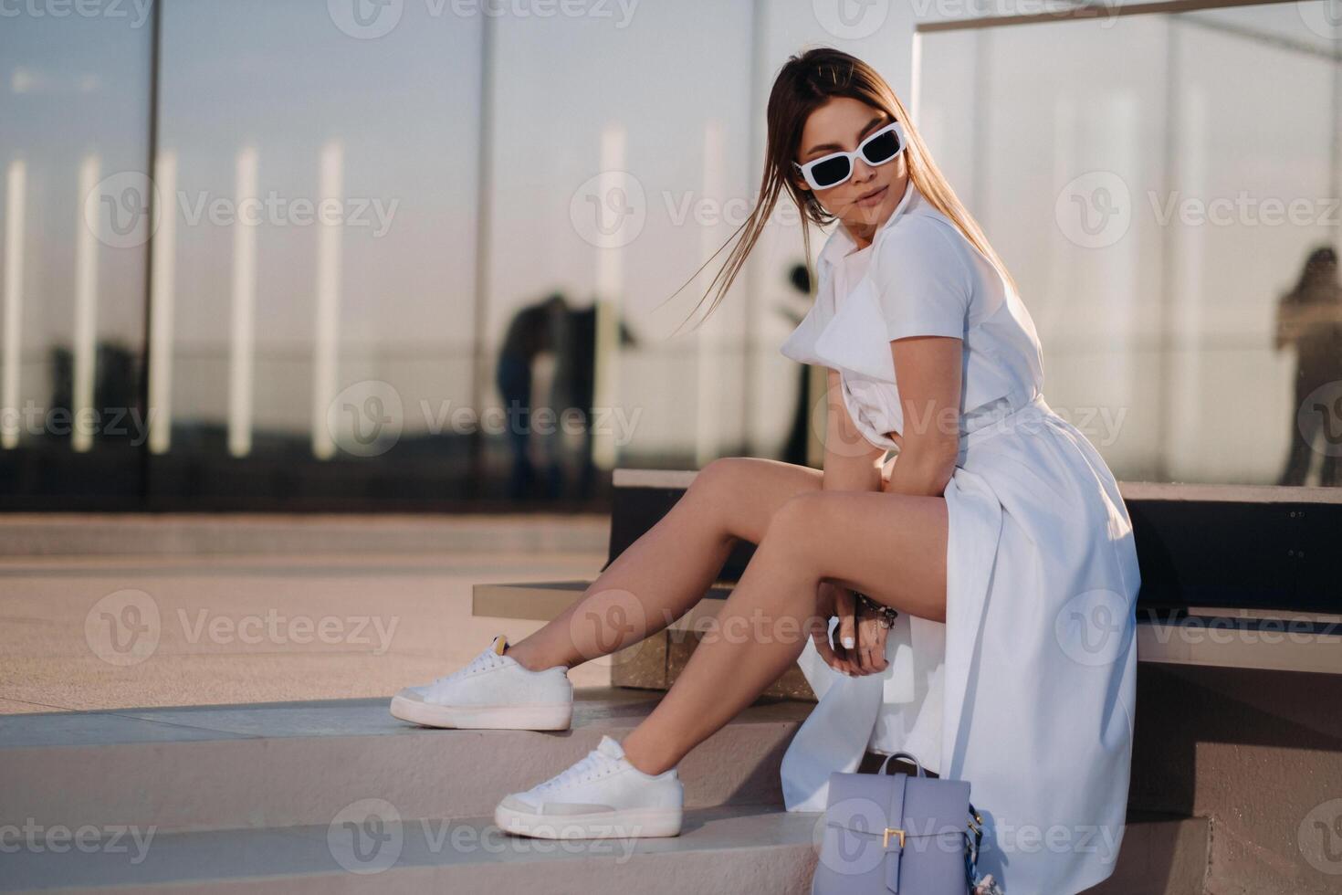 A stylish girl in glasses and a white summer coat is sitting in the city photo