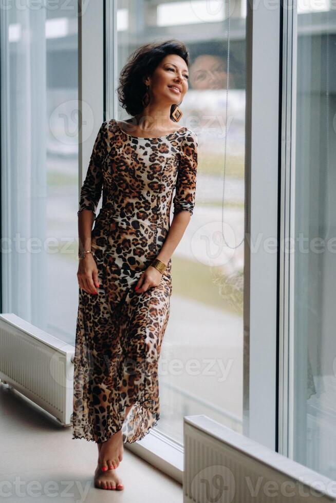 a brunette woman in a tiger dress stands near a large window photo