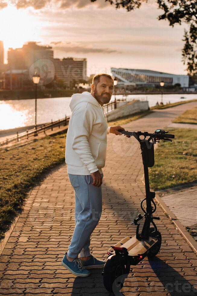 A man with a beard in white clothes rides around the city on an electric scooter at sunset photo