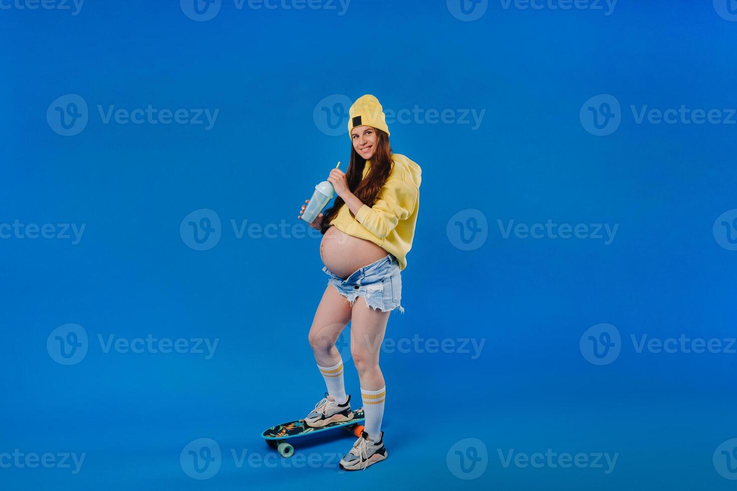 a pregnant girl in yellow clothes with a glass of juice rides a skateboard on a blue background photo