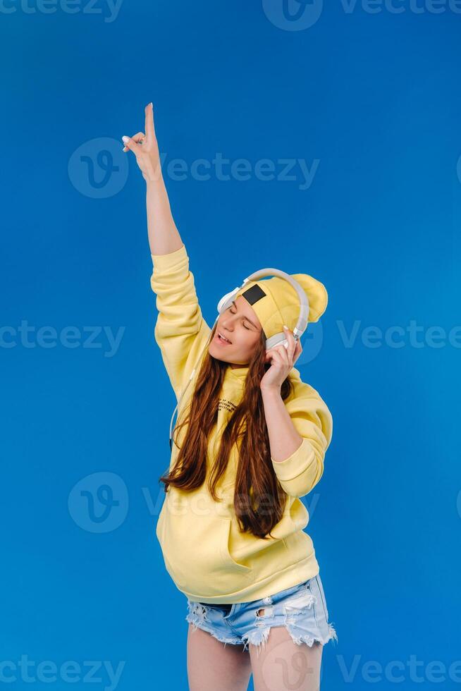 A pregnant girl in a yellow jacket and headphones stands on a blue background photo