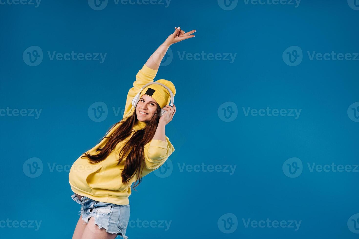 A pregnant girl in a yellow jacket and headphones stands on a blue background photo