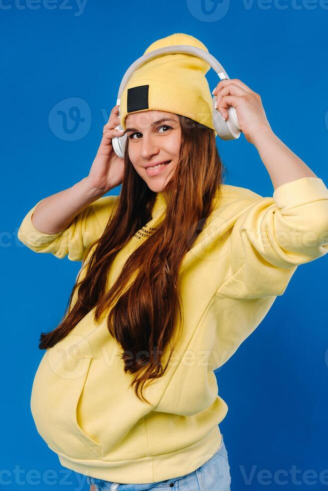 A pregnant girl in a yellow jacket and headphones stands on a blue background photo