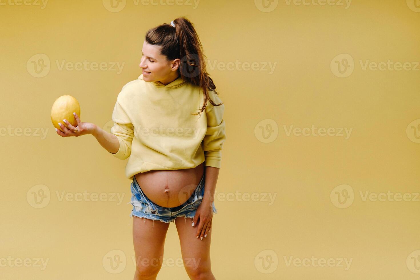 a pregnant girl with a yellow jacket stands with a melon in her hands on a yellow isolated background photo