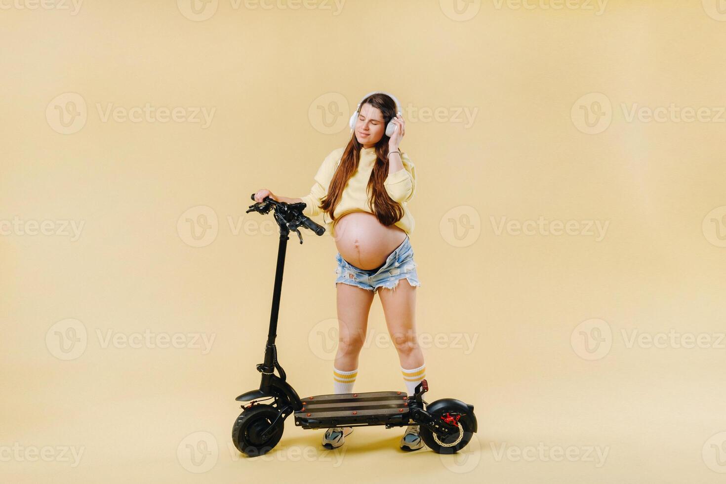 Pregnant girl in yellow clothes and headphones on an electric scooter on an isolated yellow background photo
