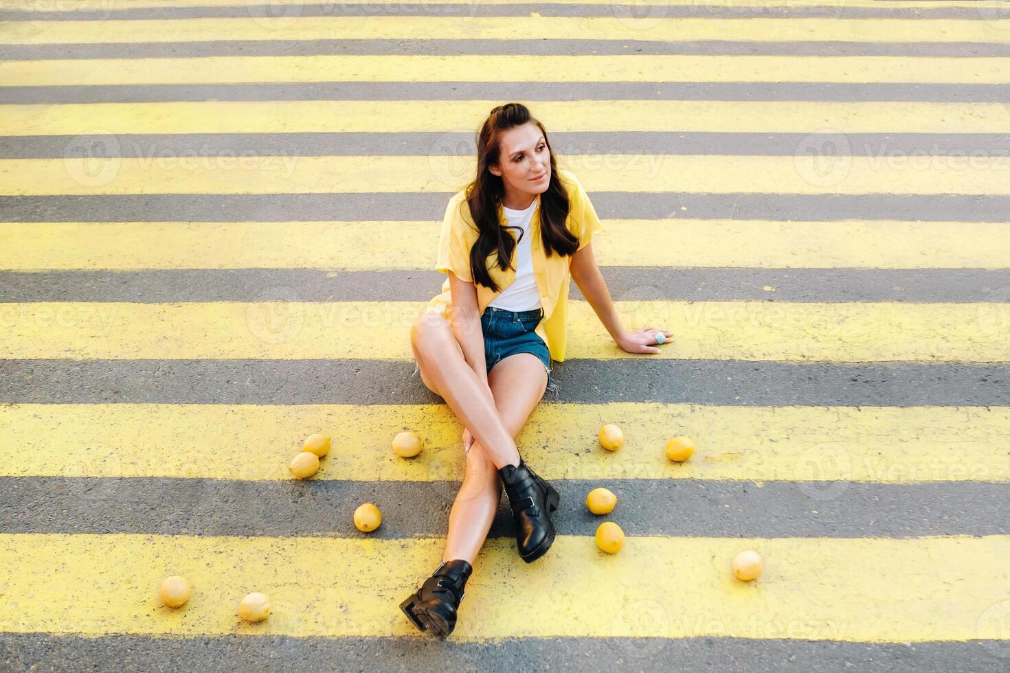 a girl with lemons in a yellow shirt, shorts and black shoes sits on a yellow pedestrian crossing in the city. The lemon mood photo