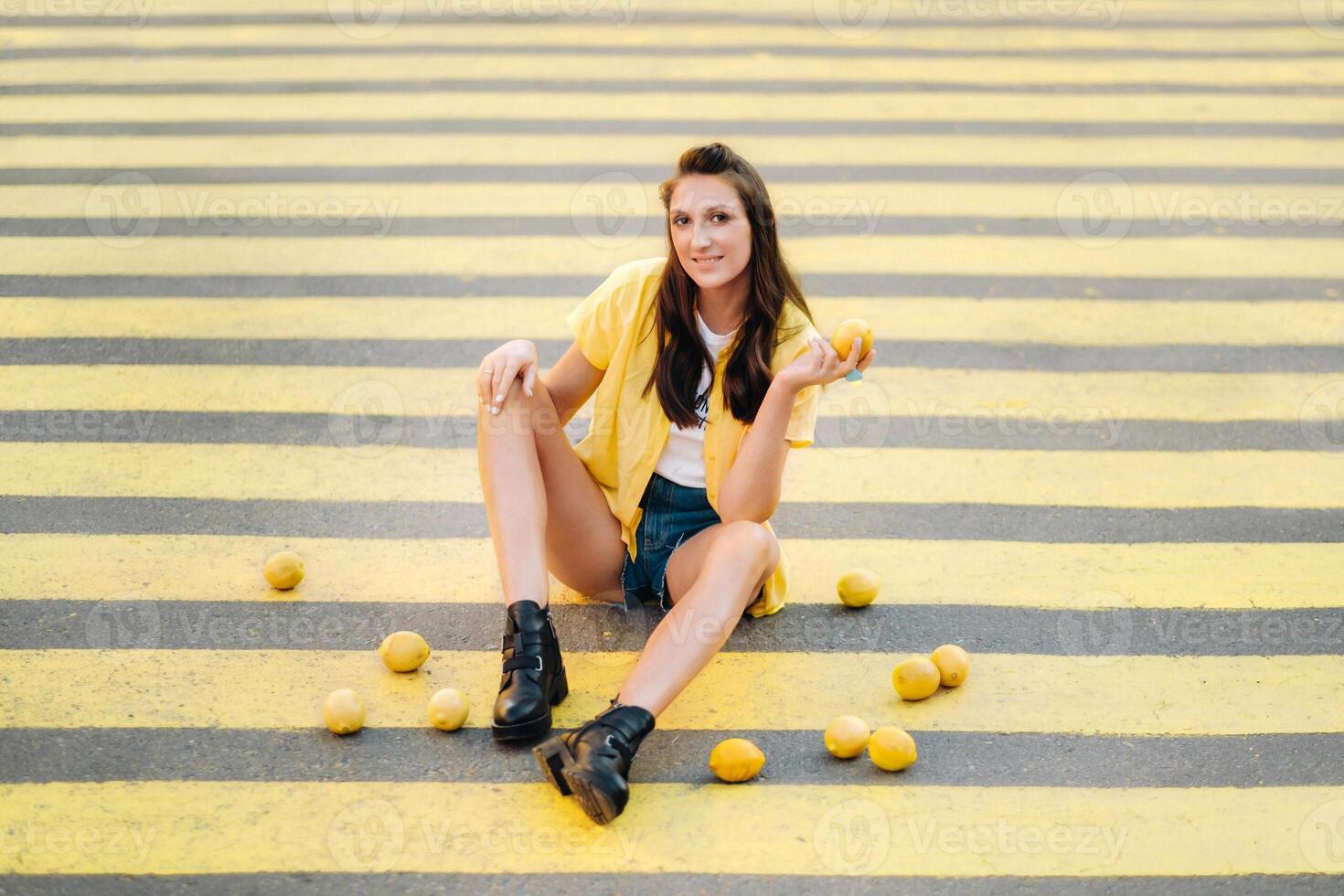 a girl with lemons in a yellow shirt, shorts and black shoes sits on a yellow pedestrian crossing in the city. The lemon mood photo