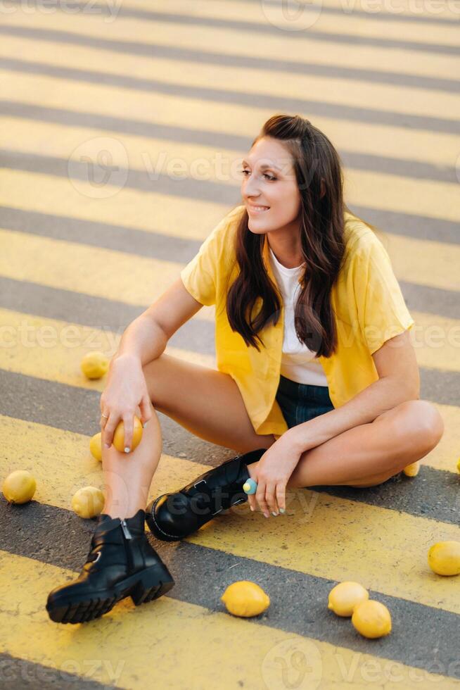 a girl with lemons in a yellow shirt, shorts and black shoes sits on a yellow pedestrian crossing in the city. The lemon mood photo