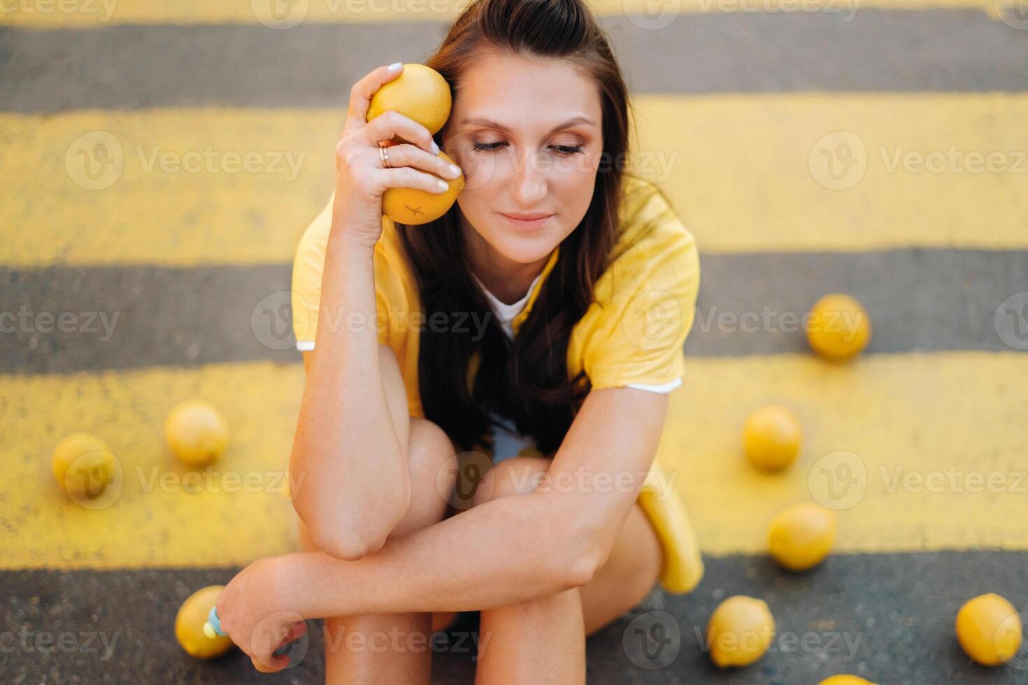 a girl with lemons in a yellow shirt, shorts and black shoes sits on a yellow pedestrian crossing in the city. The lemon mood photo