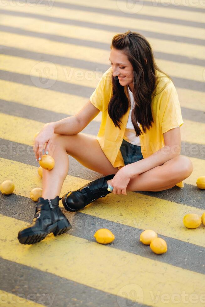a girl with lemons in a yellow shirt, shorts and black shoes sits on a yellow pedestrian crossing in the city. The lemon mood photo