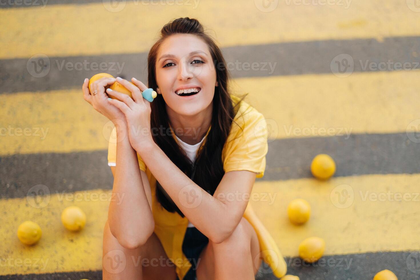 an unusual girl in a yellow shirt, shorts and black shoes with lemons is sitting on a pedestrian crossing in the city. The lemon mood. photo