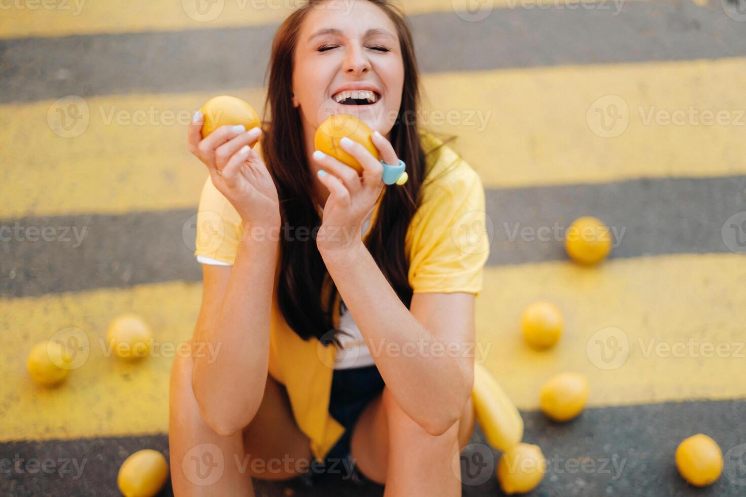a girl with lemons in a yellow shirt, shorts and black shoes sits on a yellow pedestrian crossing in the city. The lemon mood photo