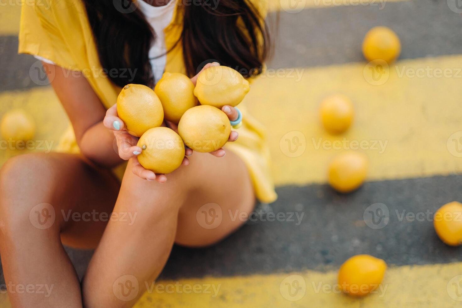 a girl with lemons in a yellow shirt, shorts and black shoes sits on a yellow pedestrian crossing in the city. The lemon mood photo