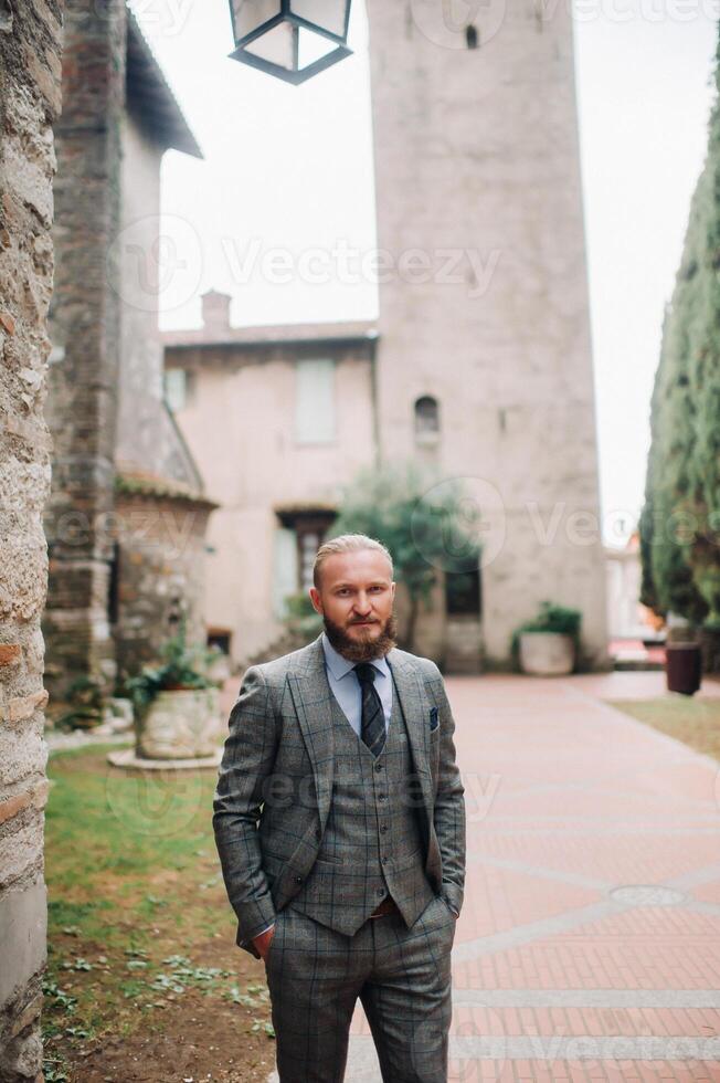 A man with a beard in a strict grey three-piece suit with a tie in the old town of Sirmione, a Stylish man in a grey suit in Italy photo