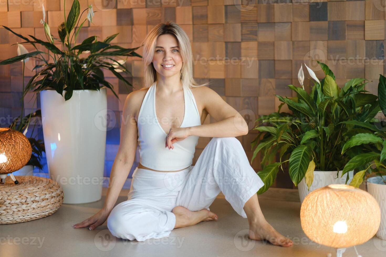 A young female yoga instructor in white athletic wear sits on the exercise floor in her studio before class photo