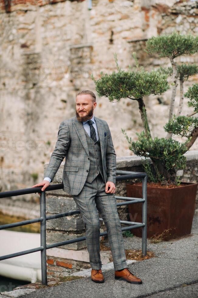 A man with a beard in a strict grey three-piece suit with a tie in the old town of Sirmione, a Stylish man in a grey suit in Italy photo