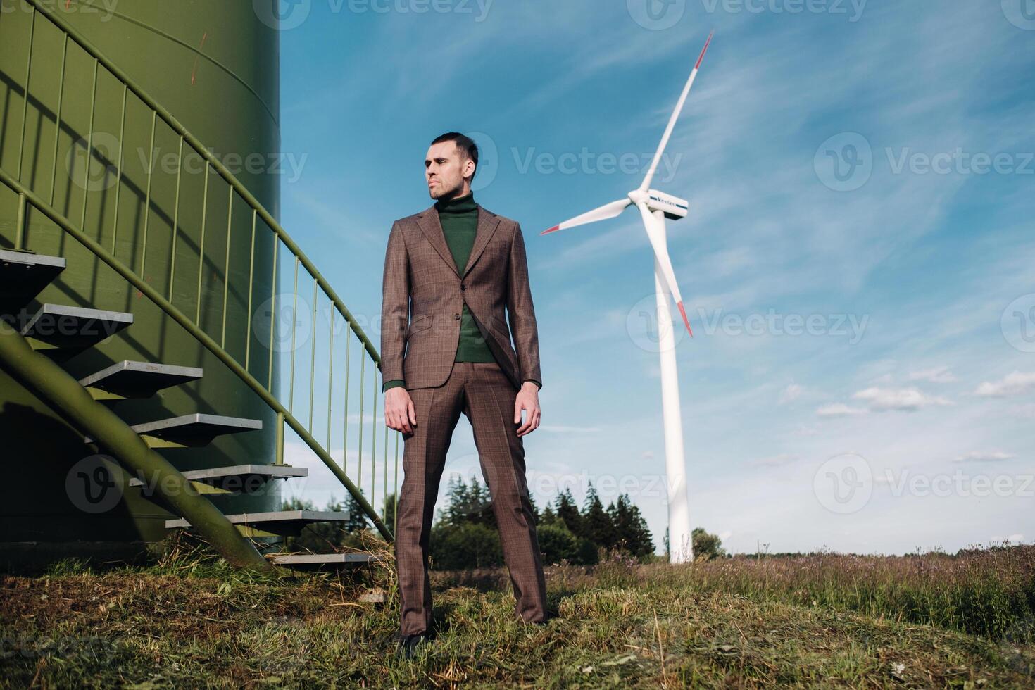 A man in a business suit with a green Golf shirt stands next to a windmill against the background of the field and the blue sky.Businessman near the windmills.Modern concept of the future. photo