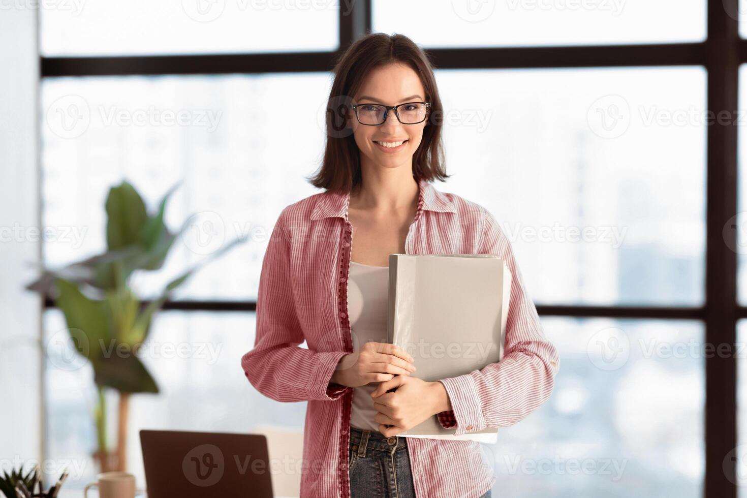 Business Career. Successful woman in glasses holding folder smiling to camera standing near window in modern office photo