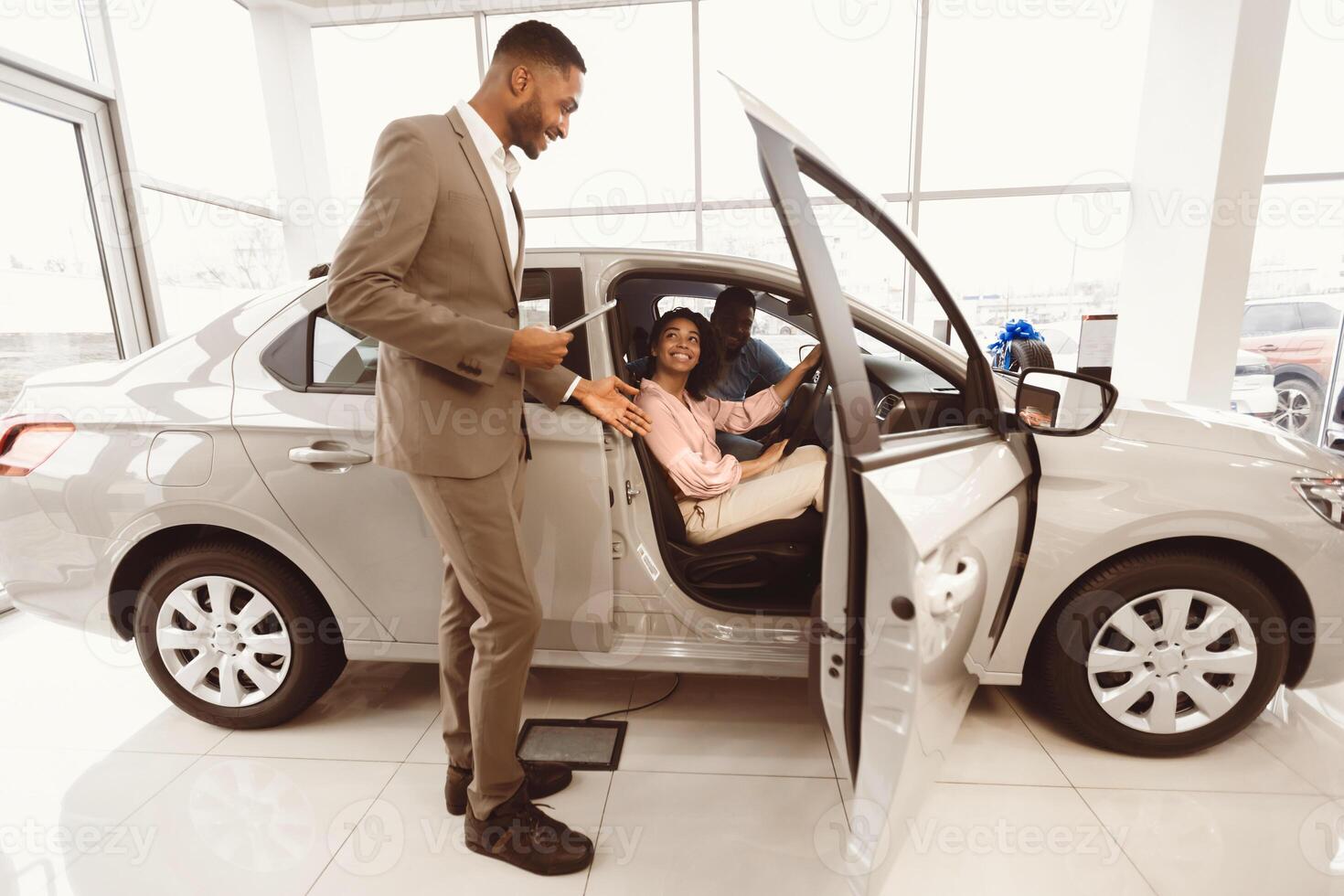 Buying Car. Cheerful Afro Spouses Sitting In Auto Testing It With Auto Sales Manager In Dealership Center photo