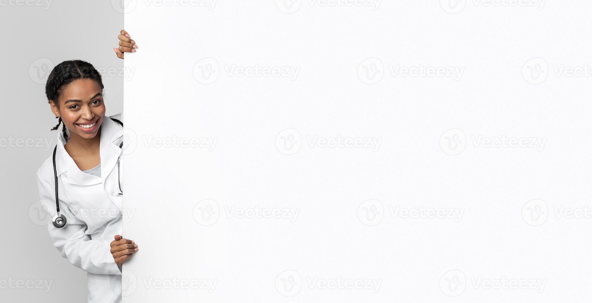 Young black female medical intern looking out behing white advertising board with free space for text and smiling, standing over light background photo
