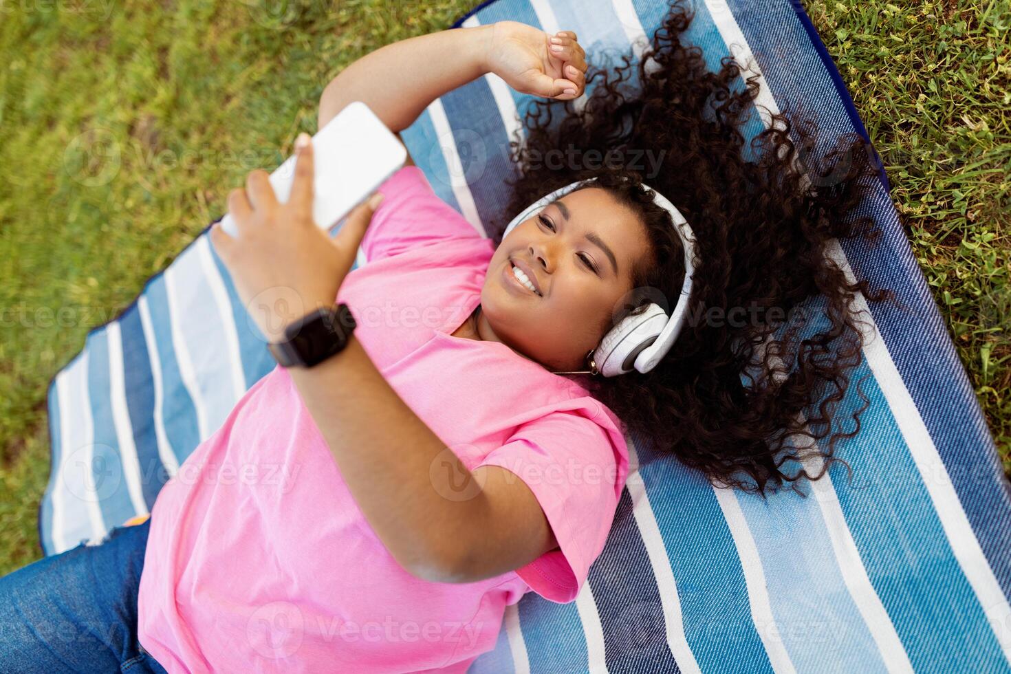 A young lady relaxes on a striped blanket in a grassy park, wearing headphones and holding a smartphone. She smiles while enjoying her music under the warm sun. photo