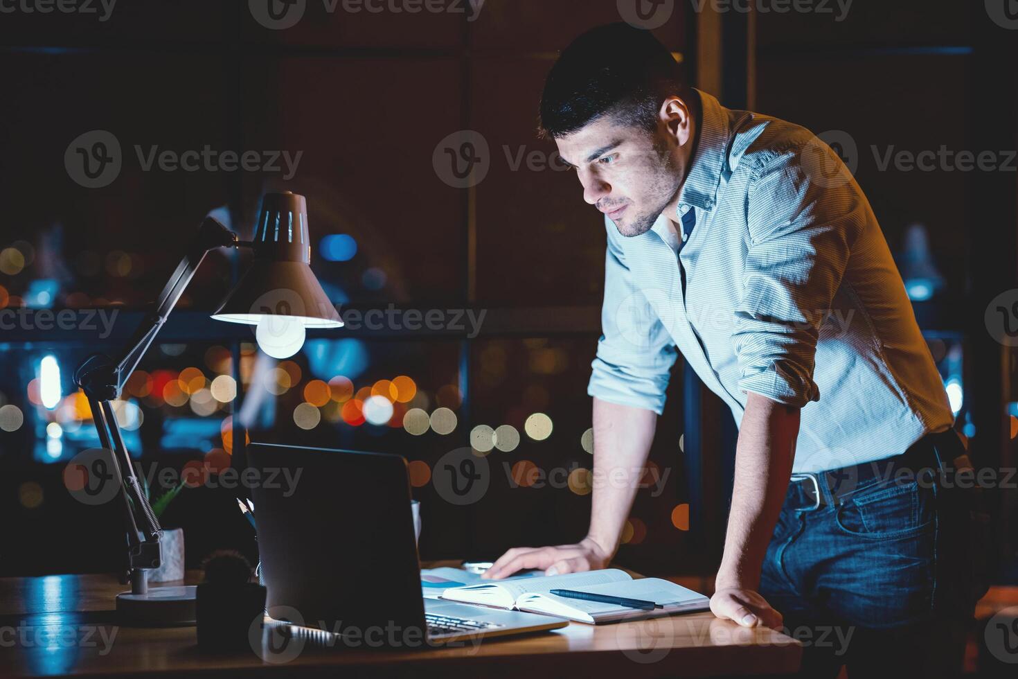 Serious Entrepreneur Man Standing Looking At Laptop Working Standing In Modern Office With Panoramic View At Night. Copy Space photo