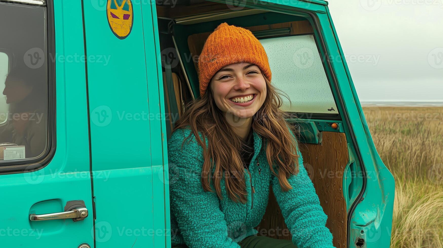 Young woman smiling in a teal vintage van during a sunny day in a grassy field, wearing an orange beanie and cozy sweater photo