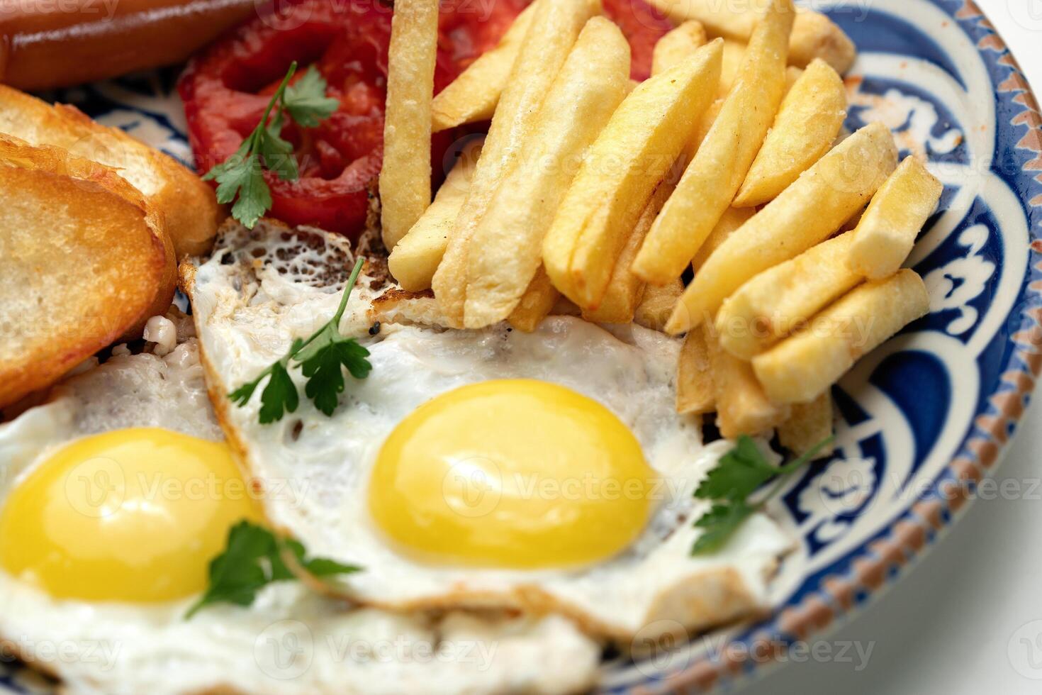 Breakfast plate with fried eggs, sausage, tomatoes, and fries served on a decorative blue plate photo