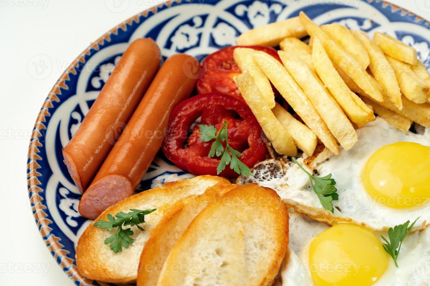 Breakfast plate with fried eggs, sausage, tomatoes, and fries served on a decorative blue plate photo