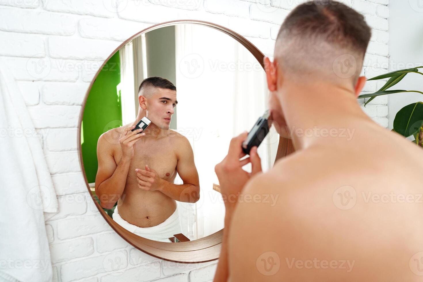 Handsome young man shaving face with electric razor on white background photo
