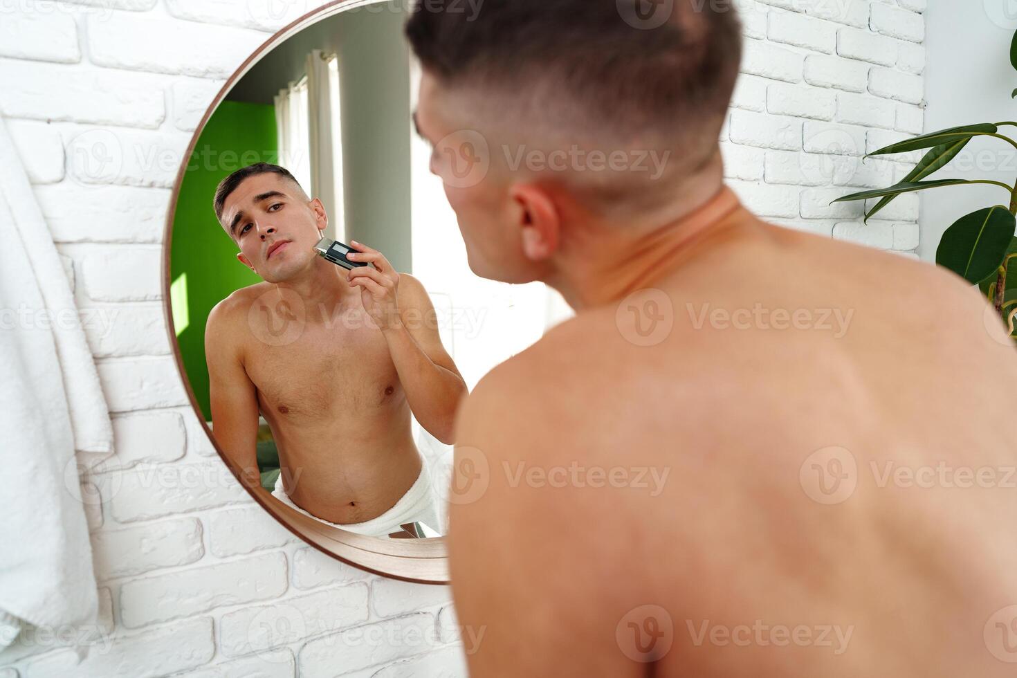 Handsome young man shaving face with electric razor on white background photo