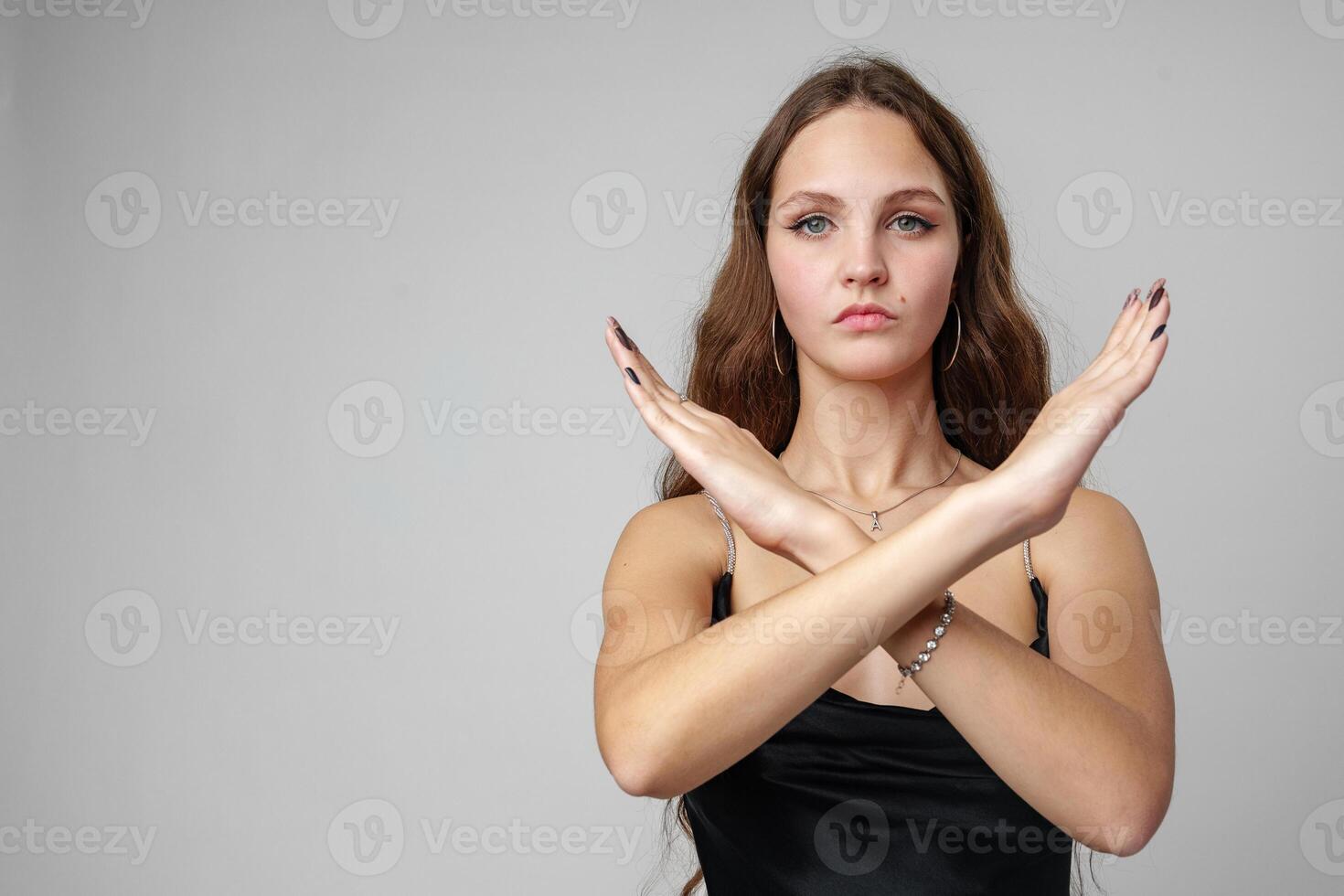 Young Woman in Elegant Black Dress Making an X Sign With Her Arms Against a Grey Background photo