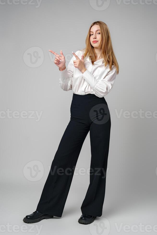 Young blonde woman in formal outfit pointing to the side against gray background photo