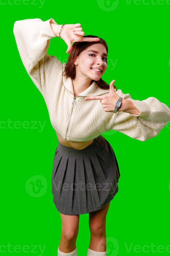 A young woman stands against a vibrant green screen, creating a finger frame with her hands as if visualizing a composition or focusing on a specific view. photo