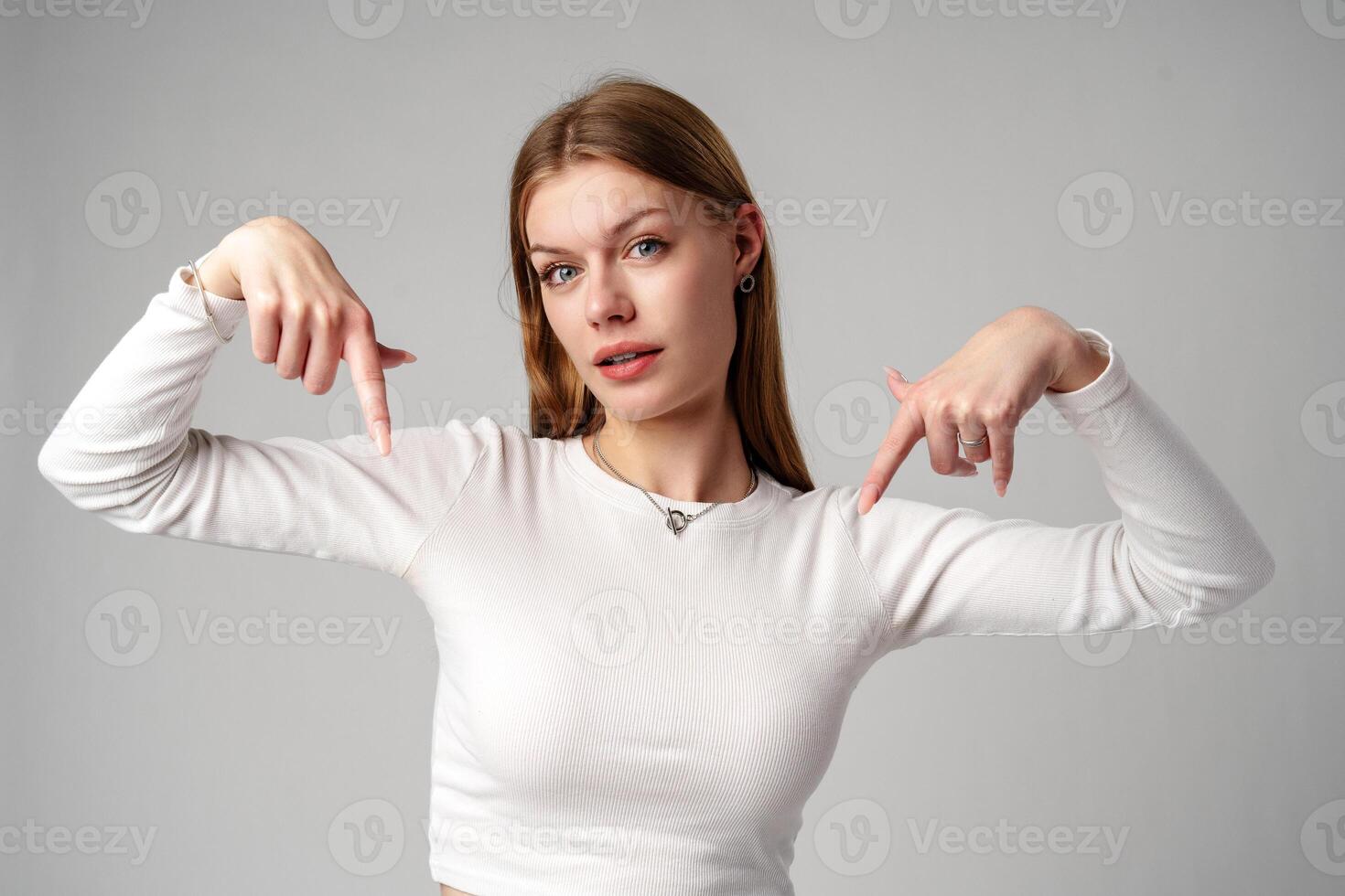 Young Woman in White Shirt Pointing Downwards With Both Hands Against a Gray Background photo
