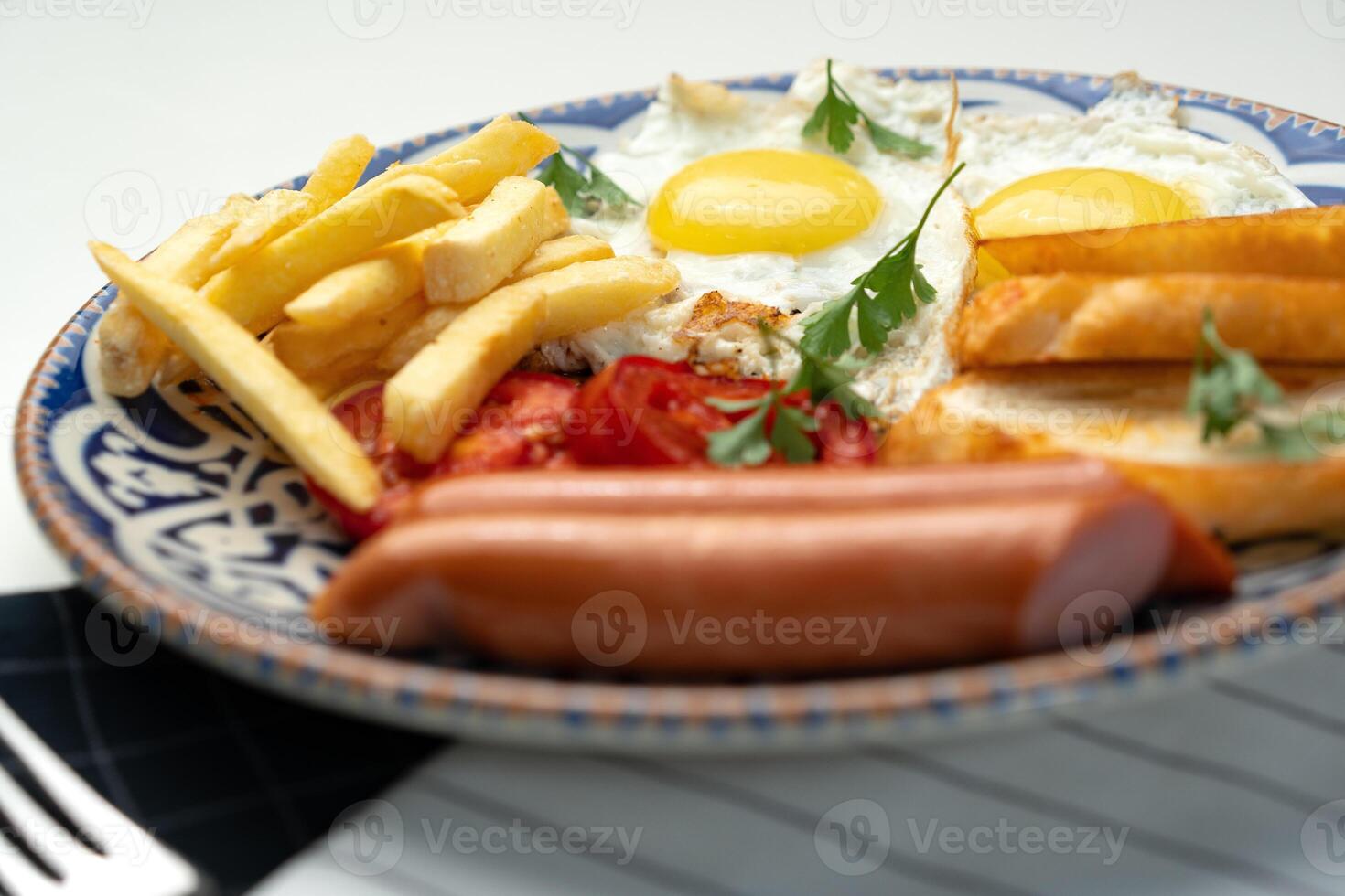 Breakfast plate with fried eggs, sausage, tomatoes, and fries served on a decorative blue plate photo