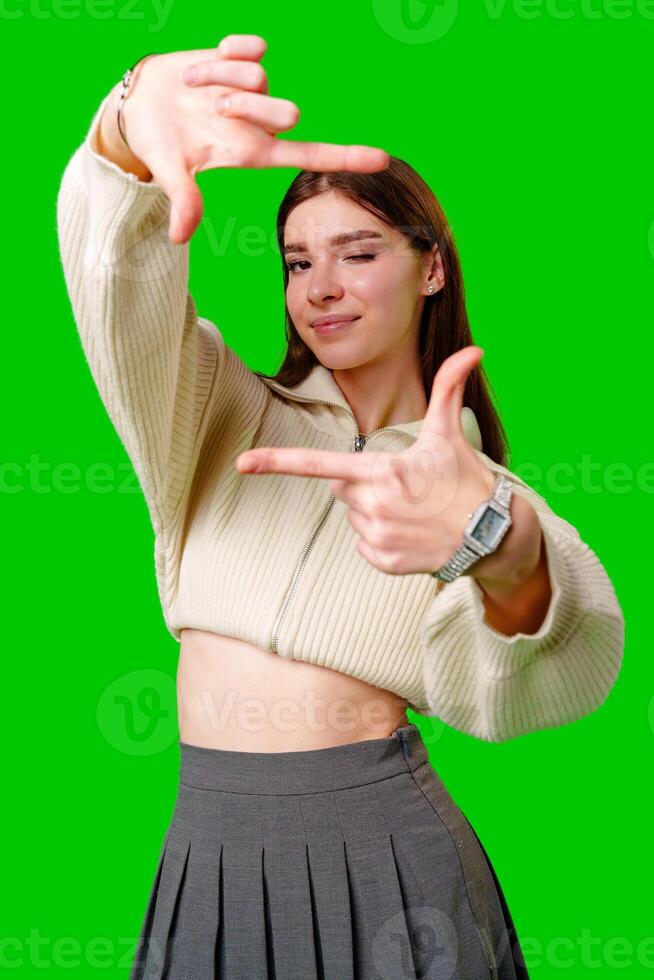 A young woman stands against a vibrant green screen, creating a finger frame with her hands as if visualizing a composition or focusing on a specific view. photo