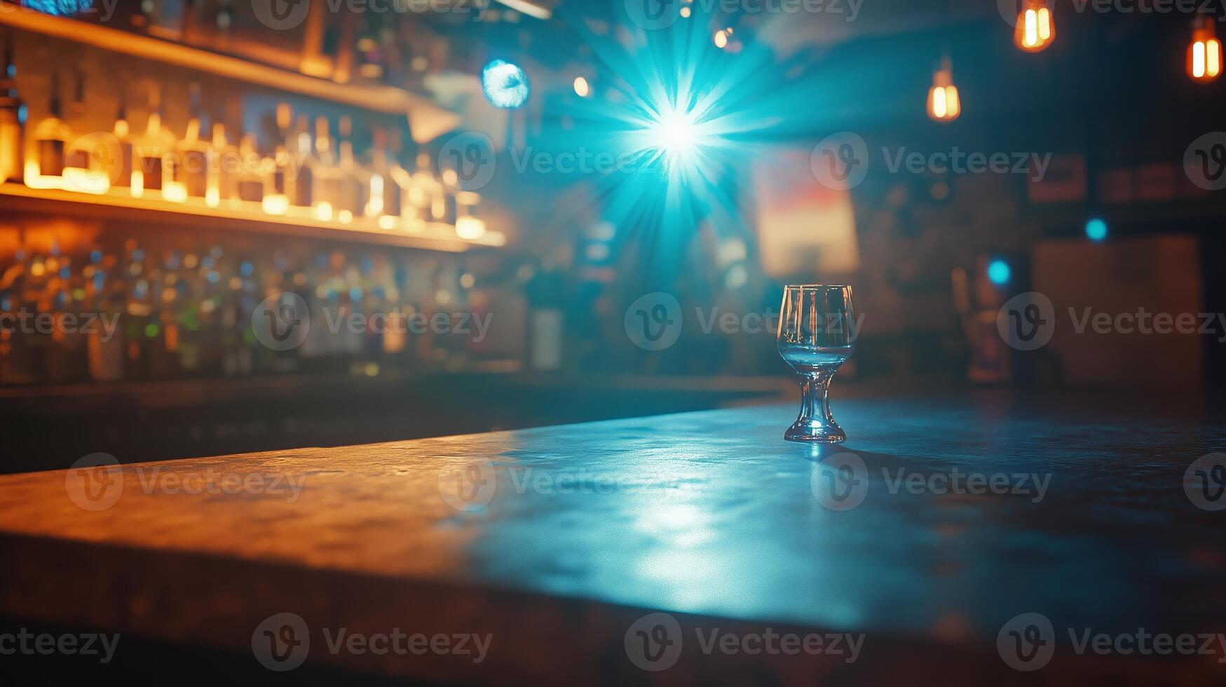 A glass sits on the bar counter in a dimly lit bar with illuminated shelves of liquor behind it during the evening photo